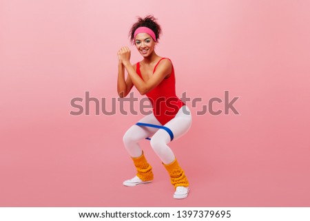 Strong african girl crouches with expander. Studio shot of active young woman training on pink background.