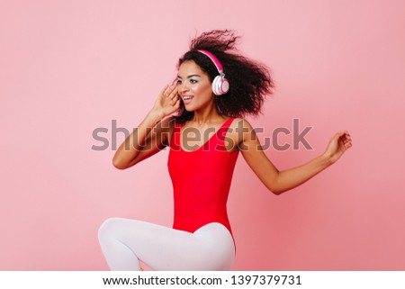 Adorable african girl in red bodysuit dancing on pink background. Curly fitness woman listening music in headphones.