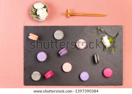 French pink, fuchsia, purple and grey  macaroons, with an incarnation white flower and other small white flowers on a slate, on the side is a pair of chopsticks, plus white incarnations flowers