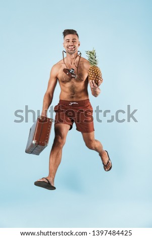 Photo of a handsome excited emotional tourist adult man posing isolated over blue wall background holding pineapple and suitcase.
