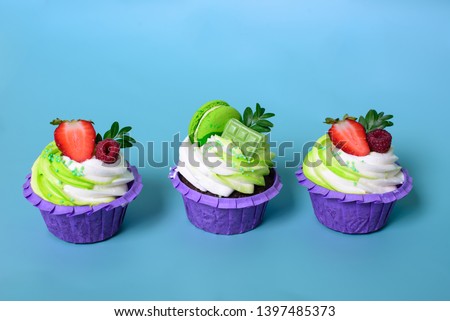 Three cupcakes with whipped cream, chocolate bar, strawberry ,decorated macaroons on blue background. Picture for a menu or a confectionery catalog. with space for text.