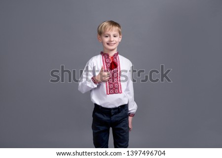 Happy smile little blond boy dressed in traditional Ukrainian clothes embroidered posing on a gray background showing hand sign all is well.
