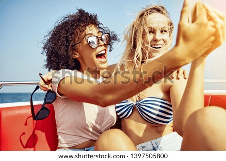 Two young female friends sitting together on a boat taking selfies together and laughing during their summer vacation