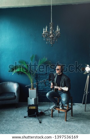 Man singer sitting on a stool in a headphones with a guitar recording a track in a home studio