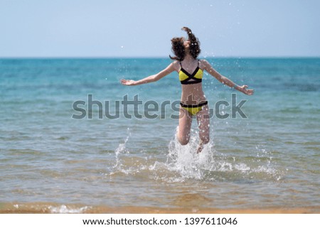 Joyful agile woman in a bikini leaping up in the ocean creating a splash of water and tossing her long hair on a sandy tropical beach