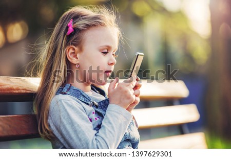 Little girl sitting in the park and having fun with smartphone. Education, lifestyle, technology concept