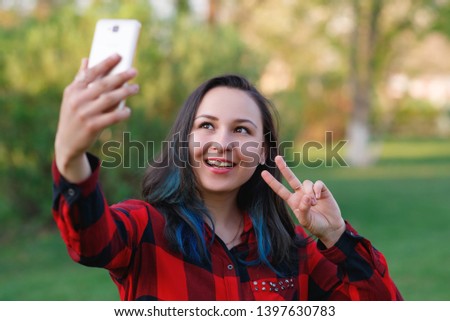 Portrait of a beautiful young woman selfie in the park with a smartphone doing v sign.