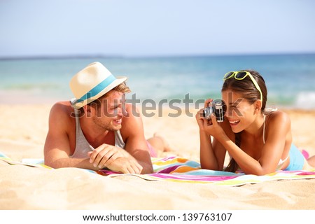 Beach fun couple travel. Woman taking photo picture of man smiling happy with retro vintage camera, Cool trendy modern hipster interracial couple on summer holidays vacation on tropical beach.