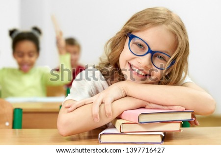 Front view of pretty female student looking at camera and laughing while lying on books in classroom. Smart girl in glasses leaning on equipment, posing and smiling at school. Concept of learning.