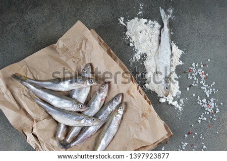 Raw smelt and flour for breading fish. One of the fish rolled in flour. View from above. Gray background. Close-up.