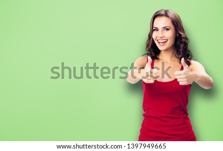 Portrait photo - young happy smiling beautiful woman in casual clothing, showing thumbs up gesture, over green color wall background. Happy girl in red dress. Brunette model at studio picture.