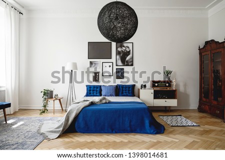 Real photo of navy blue bedroom in modern condo. Dark wooden china closet in the corner, small white dresser with vintage radio on it next to king size bed with velvet pillows and blanket.