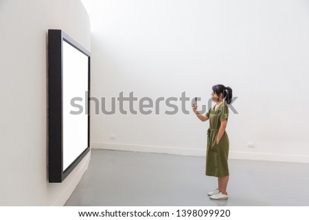 An Asian long hair woman wearing glasses in green dress shooting picture with mobile phone to white billboard in black frame in a white room.