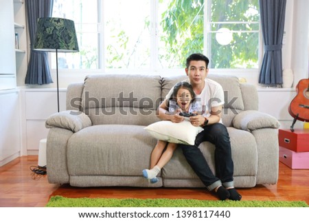 Happy Asian family, father and daughter are holding joystick and playing video game on the sofa in the living room with happy smiling face (relaxation and technology concept)