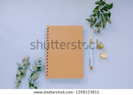 Blue desktop with eucalyptus leaves, kraft notebook, blue and gold pen and gold paperclips - neutral workspace flat lay concept
