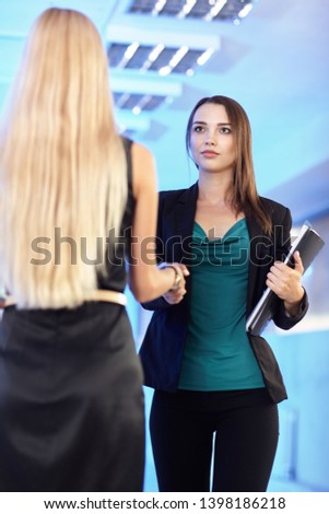 Two girls in the office with folders with papers in hand. Young women shake hands. Blue empty hall on the background