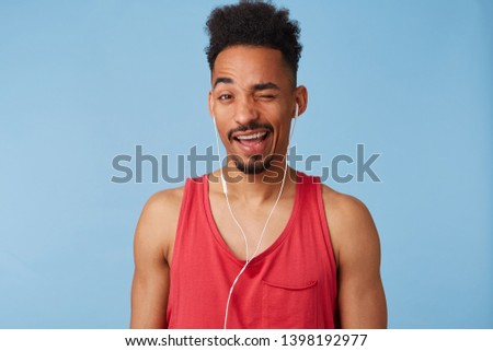Close up of young african american guy in a great mood,wears in a red jersey, listens cool music, winks and smiles broadly stands over blue background.