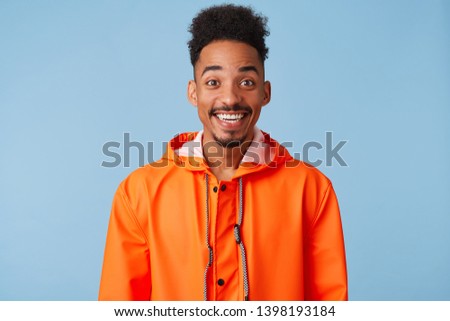 Close up of happy amazed young african american dark skinned man, feels great, wears in orange rain coat, broadly smiles isolated over blue background. Royalty-Free Stock Photo #1398193184