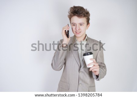young teenager with surprised emotions in grey business clothes standing with mobile phone and paper cup of coffee on white studio background