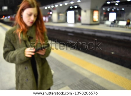 Defocused background of a young woman with long copper hair reading on a ereader near rails in a modern station. Intentionally blurred post production for bokeh effect.