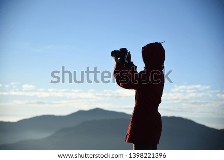 A female photographer wearing a red shirt stands shooting at the top of the blue mountain.
