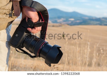 Man professional photographer with digital camera walking in tuscany fields. Photo Camera in male hands over summer landscape in Tuscany with fields, blue sky and moutains, Italy, Europe. 