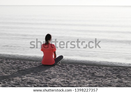 Young woman sitting on the shoreline of the sea at dawn in athletic outfit, looks towards the sea illuminated by the low sun and meditates