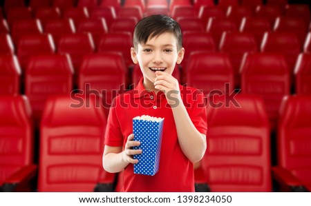 cinema, fast food and entertainment concept - portrait of smiling little boy in red polo t-shirt eating popcorn from blue paper bucket over movie theater background