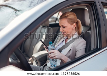 Woman taking a break from driving, using phone and drinking water while sitting in the car.