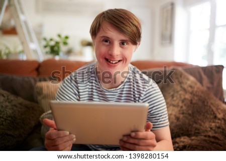 Portrait Of Young Downs Syndrome Man Sitting On Sofa Using Digital Tablet At Home Royalty-Free Stock Photo #1398280154