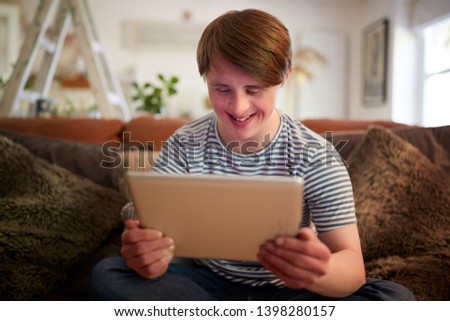 Young Downs Syndrome Man Sitting On Sofa Using Digital Tablet At Home Royalty-Free Stock Photo #1398280157