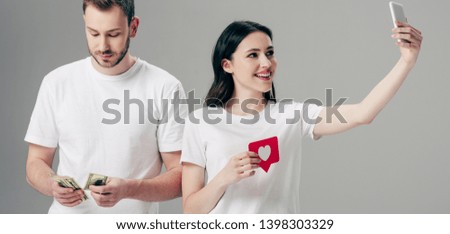 panoramic shot of smiling girl holding red paper cut card with heart symbol and making selfie with smartphone near handsome man counting dollar banknotes isolated on grey