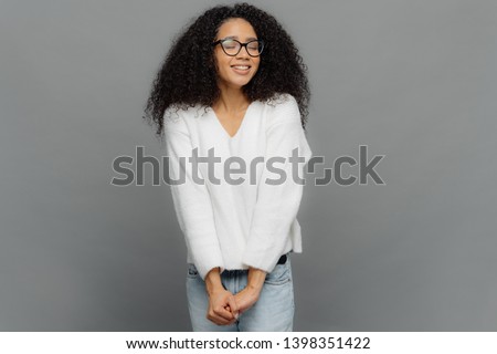 Image of happy dreamy woman keeps hands together, has eyes shut, smiles positively, has frizzy hair, wears spectacles, white jumper and jeans, isolated over grey background, Good emotions concept