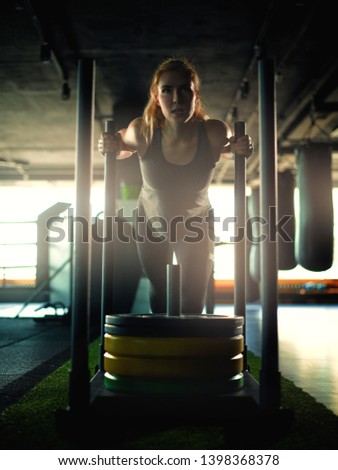 Backlit shot of determined strong young woman pulling weight sled with plates on artificial lawn grass during cross training workout in gym	
