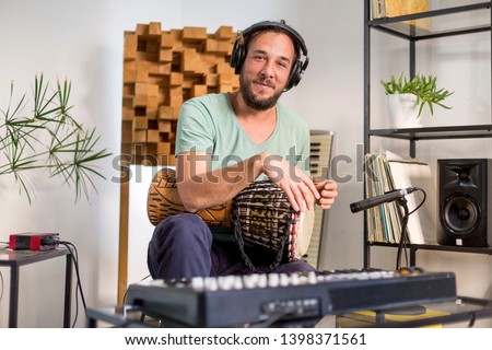 Professional musician recording djembe drum instrument in digital studio at home. He is surrounded with instruments and midi controller. Music production concept.