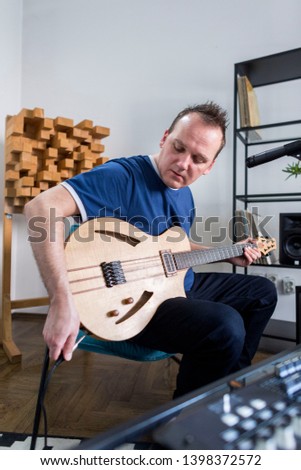 Professional musician plugging in electric guitar in digital studio at home for recording. He is surrounded with instruments and midi controller. Music production concept.
