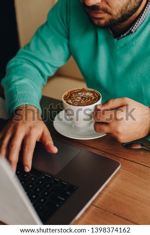 Fresh coffee for great ideas. Front view of a man working on laptop and holding cup of coffee while sitting at his working place - Image