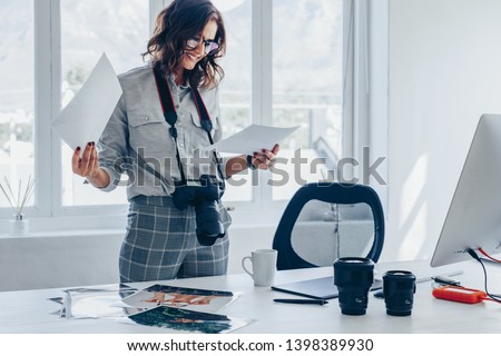 Young woman with a dslr camera around her neck standing at her desk and choosing the best images from photoshoot. Female photographer looking at the photo prints of her latest photo shoot.