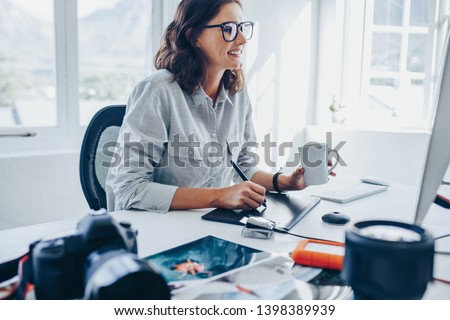 Young woman sitting at desk editing images on computer. Female photographer retouching photos in office using graphic tablet and digital pen.