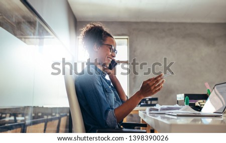 Smiling young woman talking on phone with a laptop in front. Businesswoman in conversation over phone at office. Royalty-Free Stock Photo #1398390026