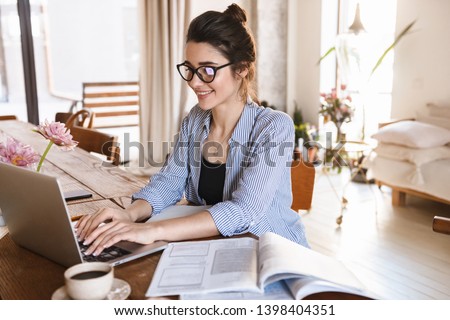 Image of smart positive woman 20s in casual clothing typing on laptop while working or studying at home Royalty-Free Stock Photo #1398404351