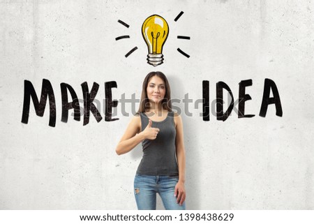 Young brunette girl wearing casual jeans and t-shirt showing thumb up with MAKE IDEA sign and cartoon light bulb drawn on white wall background. Casual clothing. Outfit ideas. Gestures and body