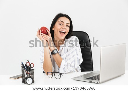Attractive young businesswoman sitting at the desk isolated over white background, holding red apple