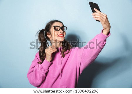 Portrait of adorable princess girl taking selfie photo on cellphone while holding paper fake sunglasses on stick isolated over blue background in studio