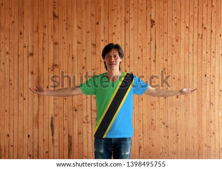 Man wearing Tanzania flag color shirt and standing with arms wide open on the wooden wall background, yellow and black diagonal band with green and blue triangle.