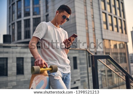 Young Caucasian American guy in glasses, on the steps with a longboard, against the background of a city building on a warm summer sunny day with a phone in his hand, listening  music with headphones.