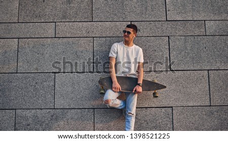 Young Caucasian American guy in glasses with a longboard against the gray wall of an urban building  listening to music with headphones and smiles.