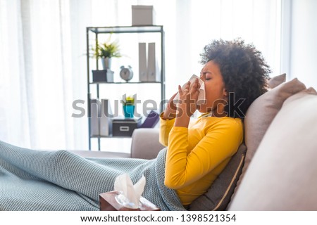 Woman sneezing with tissue while sitting on the sofa. Cold and flu.Sick woman caught cold, feeling illness and sneezing in paper wipe.Beautiful unhealthy girl covered in blanket wiping nose