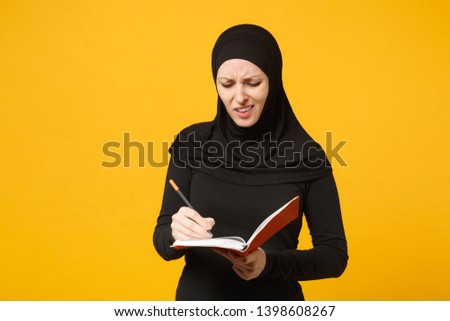 Young arabian muslim woman in hijab black clothes hold notebook, writing, prepare to exam isolated on yellow wall background, studio portrait. People religious lifestyle concept. Mock up copy space