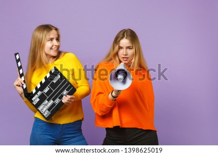 Two beautiful young blonde twins sisters girls hold classic black film making clapperboard, scream on megaphone isolated on violet blue background. People family lifestyle concept. Mock up copy space